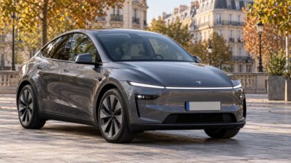 Tesla Model Y gris vu de trois quarts avant, stationné sur une place pavée bordée d’arbres aux couleurs d’automne.